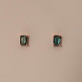 A pair of emerald cut sapphire stud earrings set in 9ct rose gold, displayed against a pink background.
