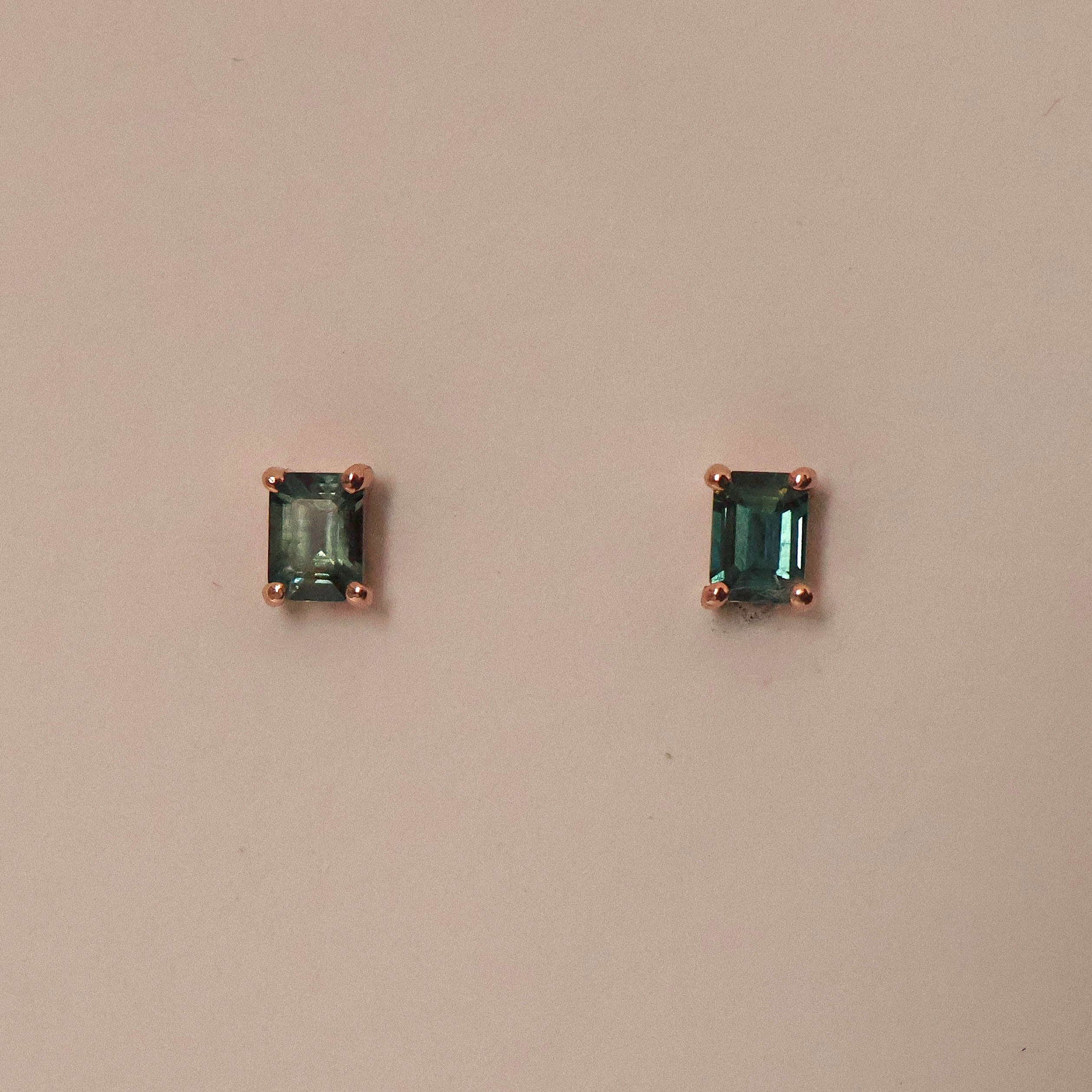 A pair of emerald cut sapphire stud earrings set in 9ct rose gold, displayed against a pink background.