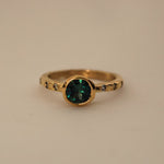 An 18ct yellow gold ring with an organic textured band, featuring a central bezel set with a teal Australian sapphire, surrounded by six light green Australian sapphire accents.