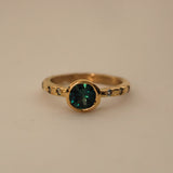 An 18ct yellow gold ring with an organic textured band, featuring a central bezel set with a teal Australian sapphire, surrounded by six light green Australian sapphire accents.