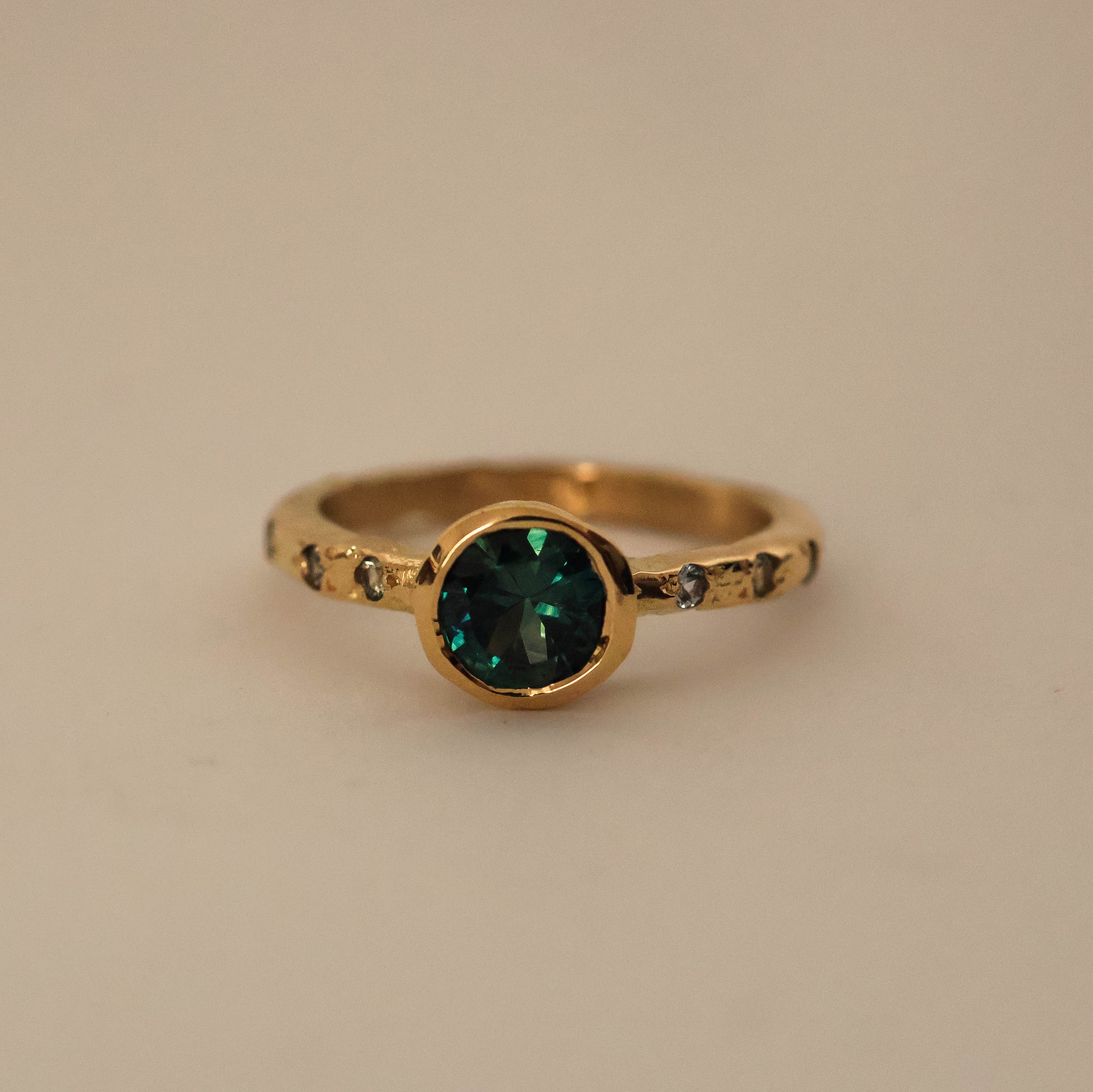 An 18ct yellow gold ring with an organic textured band, featuring a central bezel set with a teal Australian sapphire, surrounded by six light green Australian sapphire accents.