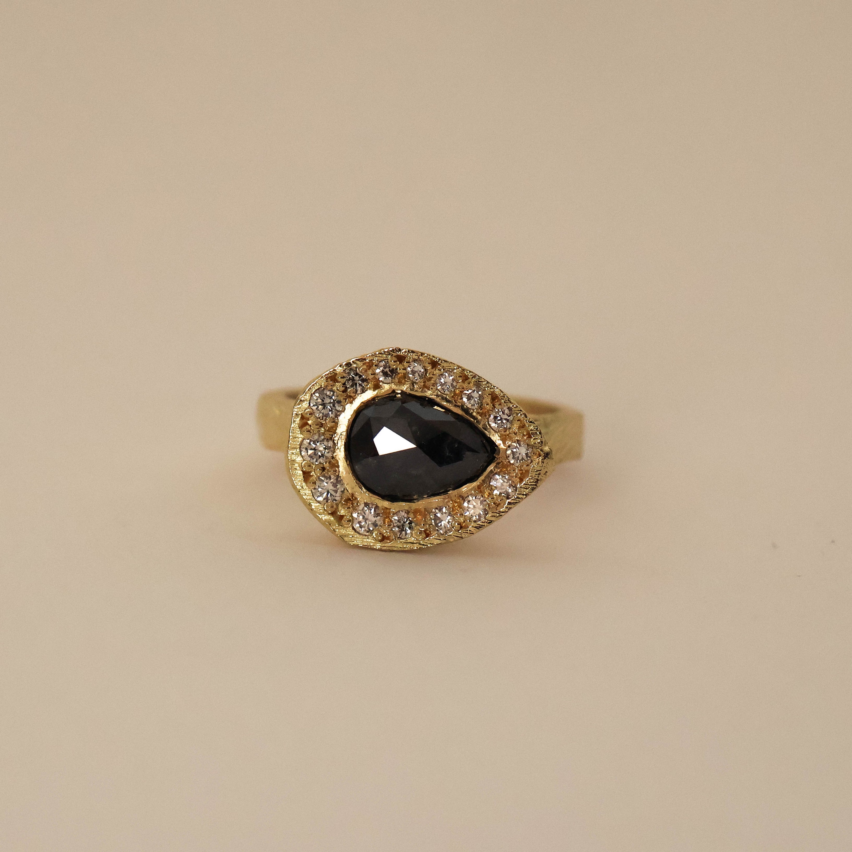 An 18ct yellow gold ring with a pear shaped salt and pepper diamond centerpiece and round conflict-free white diamonds set around it.