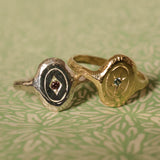 Two textured signet-style rings, one silver-toned with a diamond shape set small red gem, and one gold-toned with a light blue gem in a star setting, displayed on a green patterned surface.