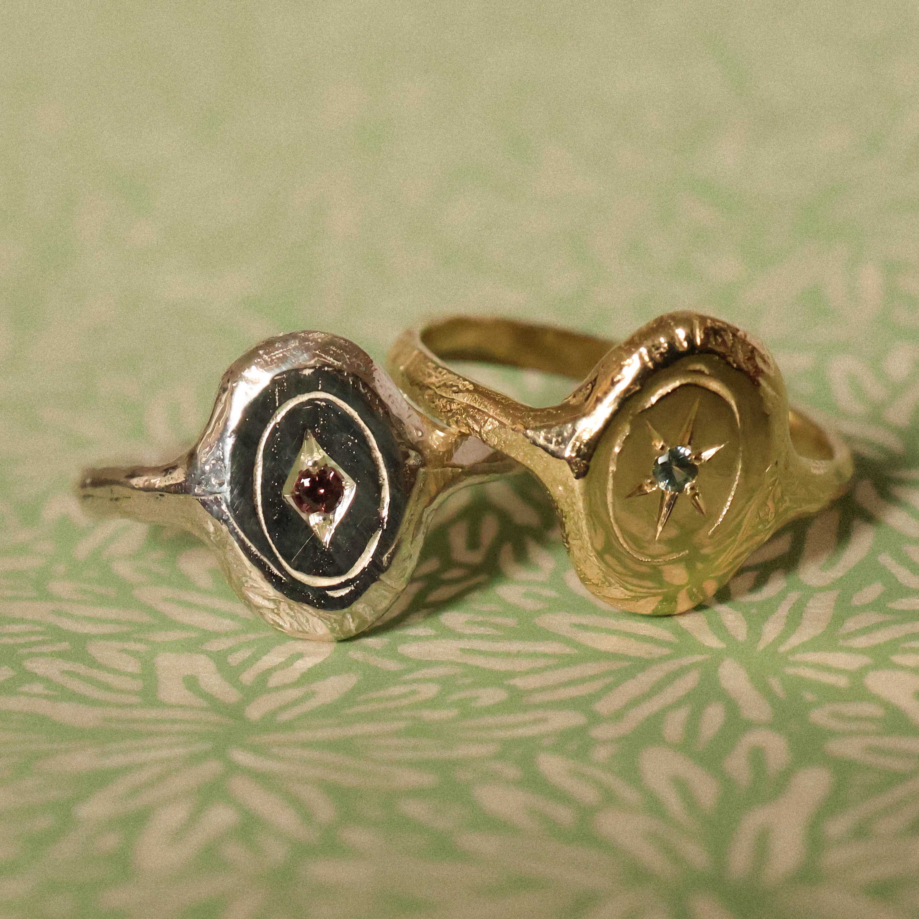 Two textured signet-style rings, one silver-toned with a diamond shape set small red gem, and one gold-toned with a light blue gem in a star setting, displayed on a green patterned surface.
